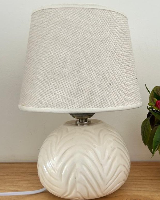 Lámpara de base esférica en cerámica blanca con relieve de hojas y pantalla de lino crudo, junto a un florero